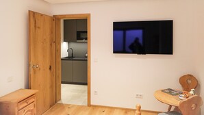 A modern room with a wooden door, a TV mounted on the wall, a small wooden table, and a kitchen visible through an open door.