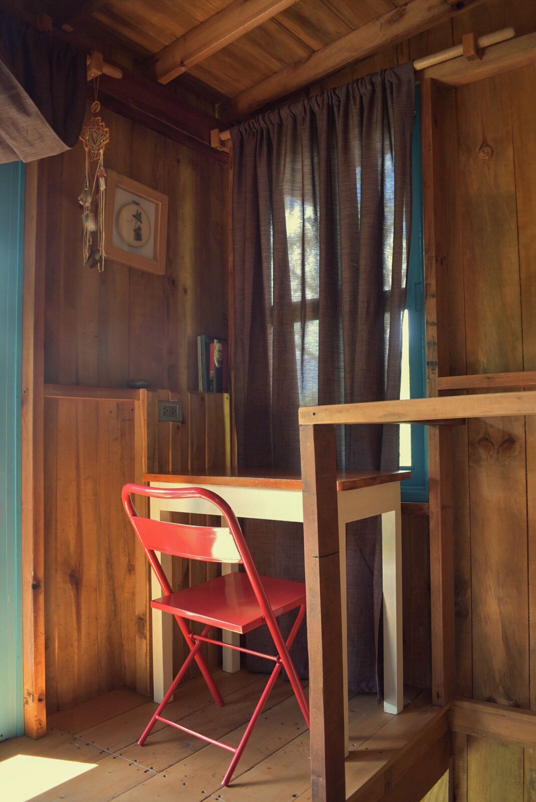 A wooden cabin interior with a red folding chair, a table, and a window with curtains.
