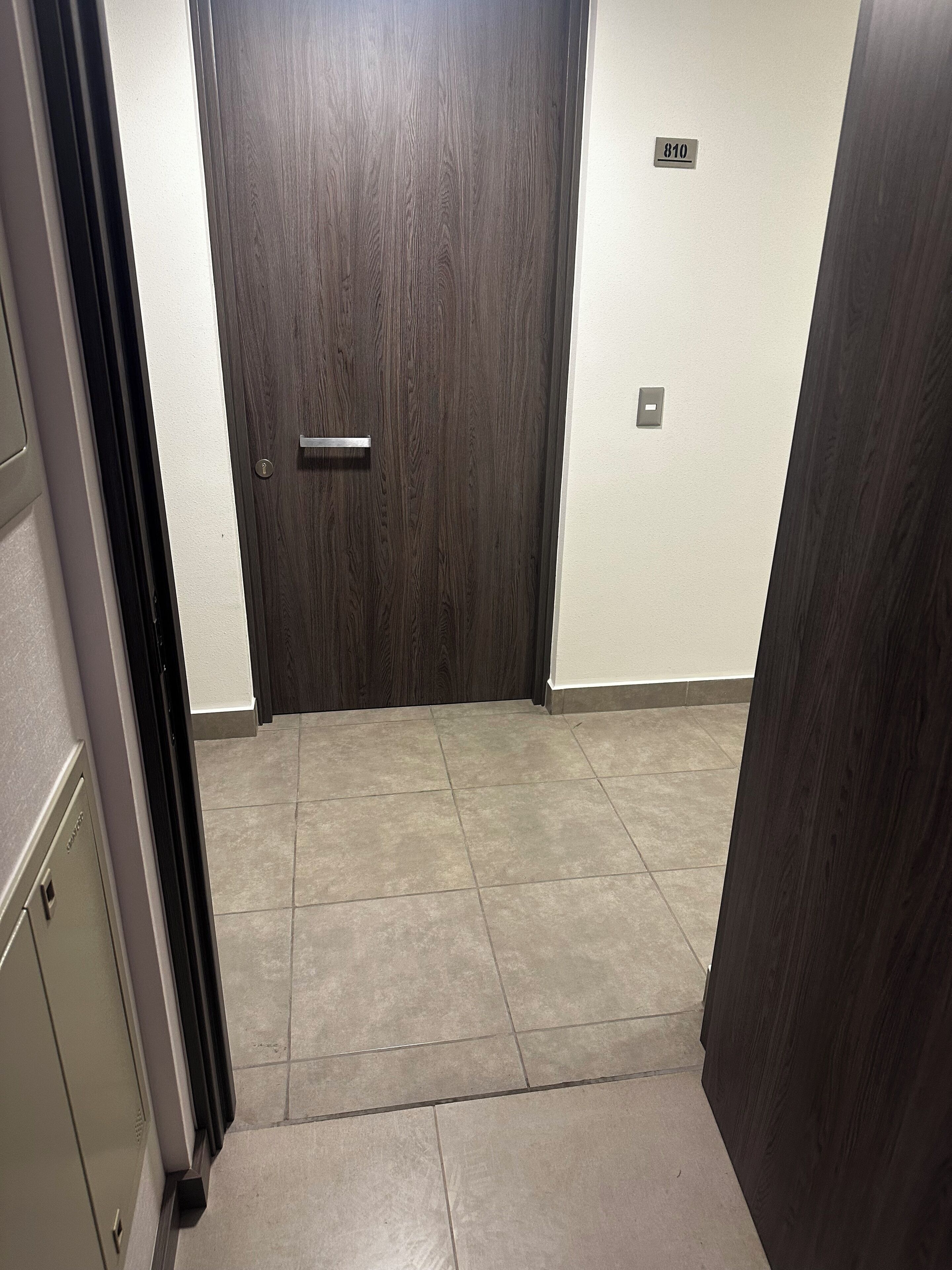 Un couloir avec une porte en bois, un interrupteur et une plaque numérotée sur le mur.