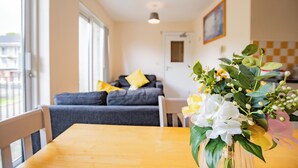 Ein Essbereich mit einem Holztisch, einer Vase mit Blumen, einem grauen Sofa mit gelben Kissen und einer karierten Wand im Hintergrund.