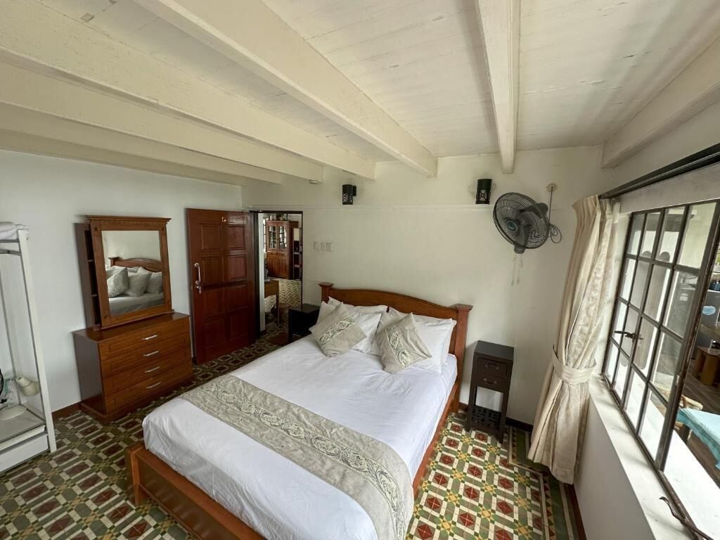 Quarto com cama, cómoda, espelho, guarda-roupa, mesa de cabeceira, ventoinha e janela com cortinas.