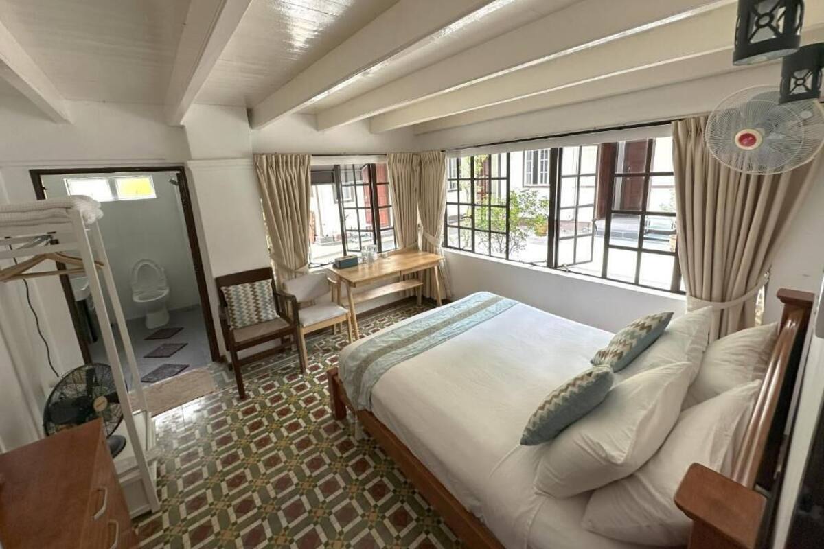 Une chambre avec un lit, un bureau, une chaise, un ventilateur et une vue sur l’extérieur.