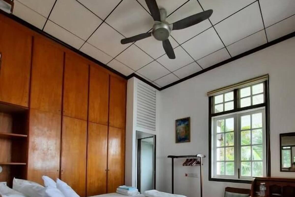 Une pièce comprenant un lit, une armoire en bois, un ventilateur de plafond, une fenêtre et une chaise.