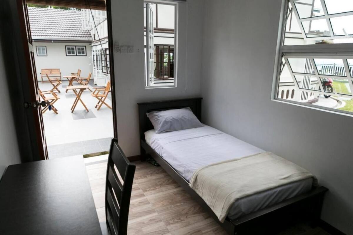 Une petite chambre avec un lit simple, un bureau en bois, deux fenêtres et une vue sur l’extérieur.