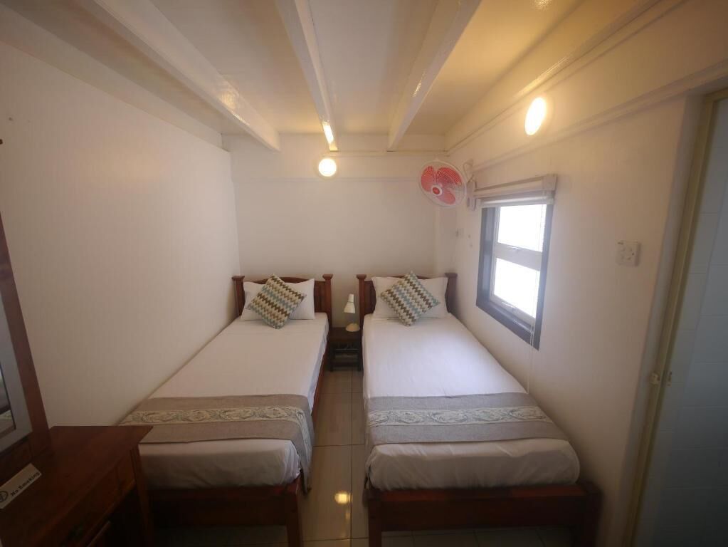 A room with two single beds, a small table, a window with a view, and a ceiling fan.