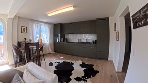 Een moderne woonkamer met een bank, een koehuidkleed en een keuken met donkere kastjes en een raam.