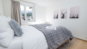 Een slaapkamer met een bed, een raam met gordijnen, een grijze deken en drie herten schilderijen aan de muur.