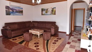 Ein Wohnzimmer mit einer Eckcouch, einem Couchtisch und einem gemusterten Teppich. An den Wänden hängen zwei Gemälde, eine Chandelier und die Decke ist aus Holz.