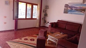 Ein Wohnzimmer mit einem roten Sofa, einem Holztisch, einem gemusterten Teppich, einem Fenster mit Vorhängen und einer Wand-Klimaanlage.