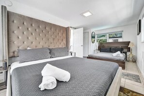 Une chambre avec un grand lit, un lit plus petit, une tête de lit capitonnée, une table de chevet, une lampe et une vue sur la verdure à l’extérieur.