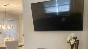 Moderni olohuone, jossa seinälle on asennettu taulutelevisio, tumma puinen konsolipöytä, jonka päällä on koriste-esineitä, sekä ruokailutila, jossa on valkoiset tuolit ja kattokruunu.