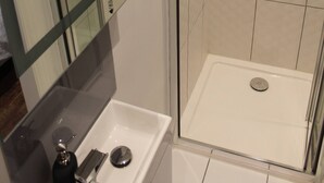 Ducha con cerramiento de vidrio, un lavabo blanco con dispensador de jabón y un espejo con marco de madera.