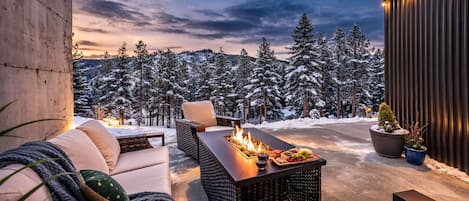 Un espace extérieur aménagé avec des sièges et un foyer, entouré d’arbres couverts de neige et offrant une vue sur le ciel.