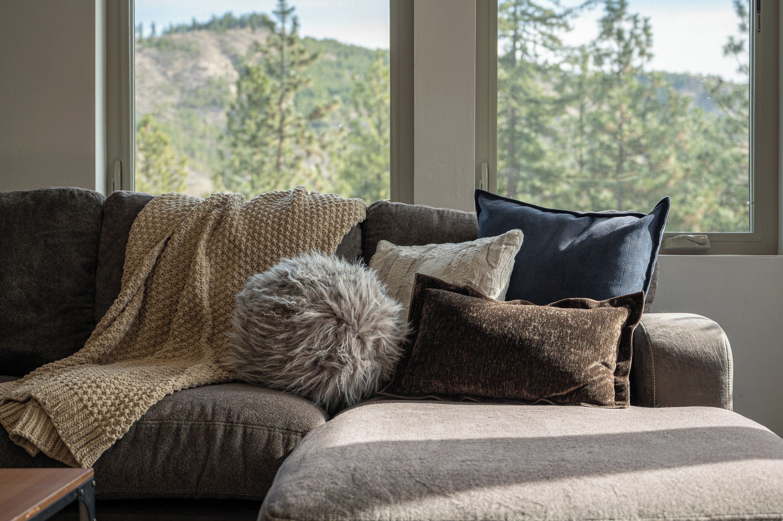 A sofa with a knitted blanket and pillows, a fluffy throw, and a view of a forest through large windows.