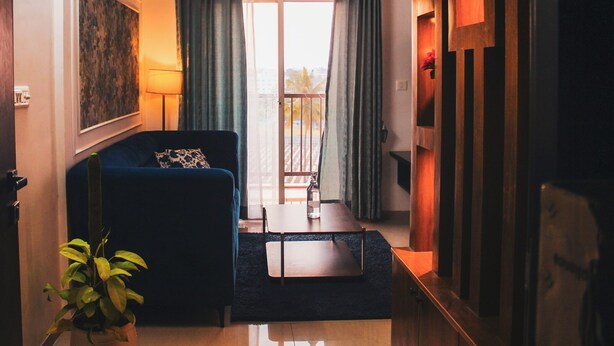 Une pièce comprenant un canapé, une petite table, une plante en pot, un tableau accroché au mur et une vue par la fenêtre, laquelle est dotée de rideaux.