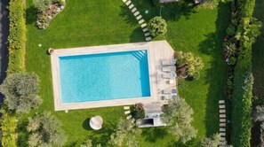 An aerial view of a rectangular swimming pool with a wooden deck, surrounded by well-maintained grass and trees.
