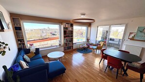 Ein geräumiges Wohnzimmer mit einem blauen Sofa, einem Esstisch mit Stühlen und großen Fenstern mit Blick auf die Stadt.