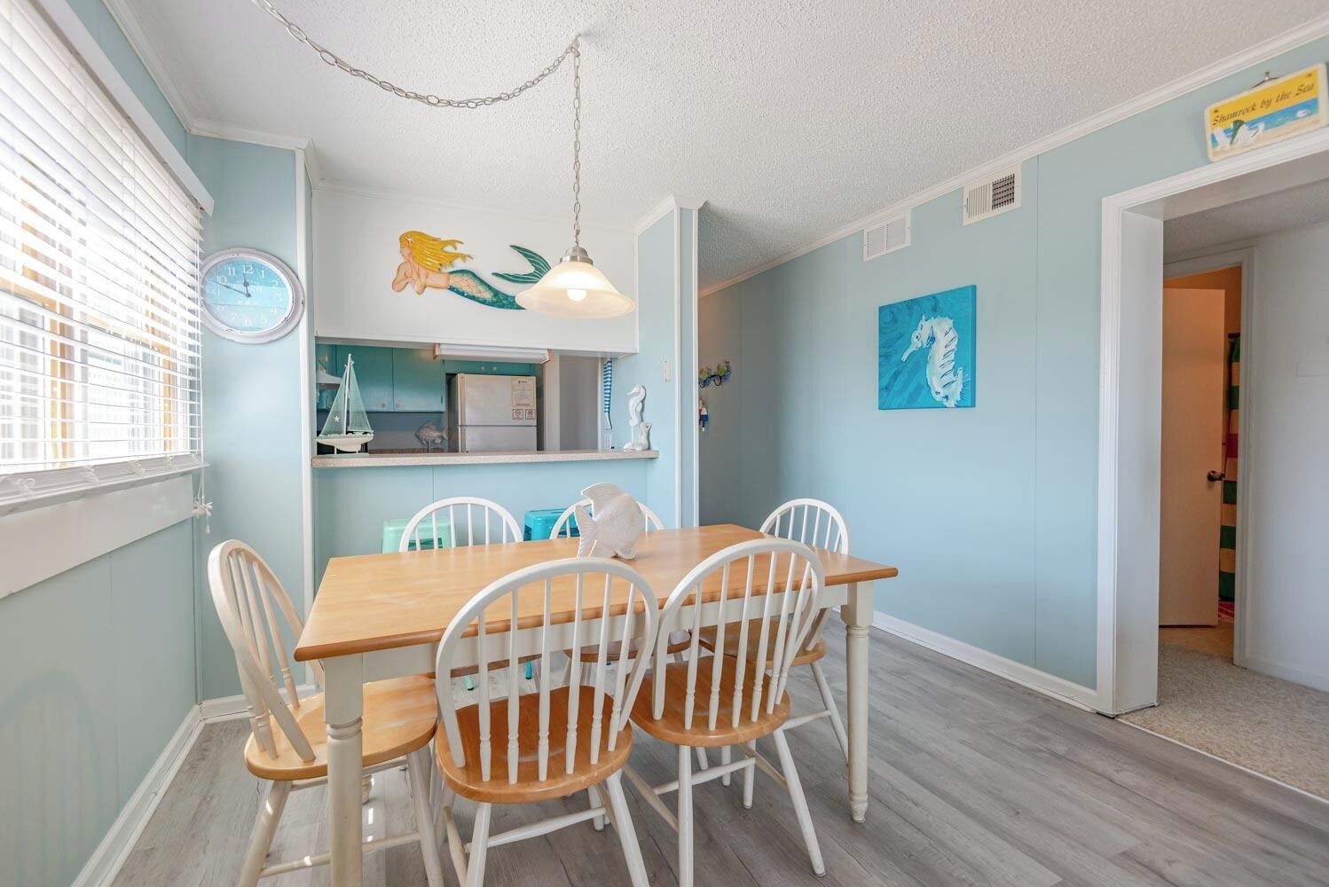 A dining area with a wooden table and white chairs, a wall clock, and a seahorse painting.