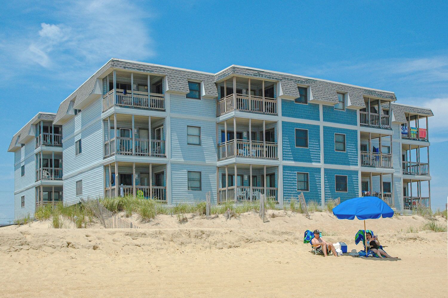 A beachfront building with multiple balconies, a sandy beach in front, and a blue umbrella.