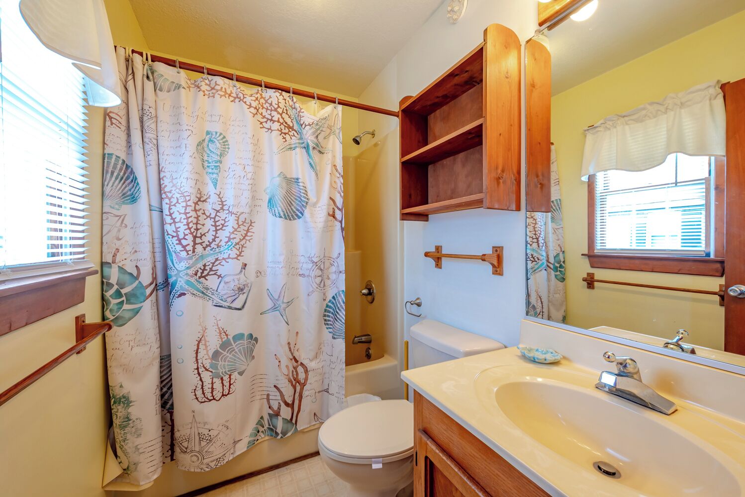 A bathroom with a shower curtain featuring sea life, a toilet, a sink with a mirror, and wooden shelves.