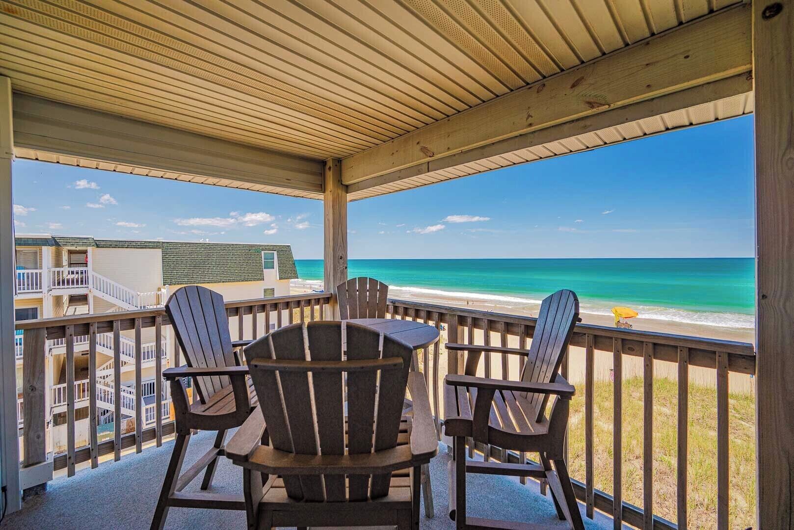 A covered outdoor seating area with Adirondack chairs and a table, overlooking a beach and ocean.
