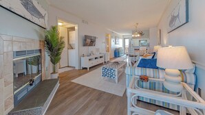 A living room with a fireplace, a sofa, a dining area, and a kitchen in the background. - JL #R-10 | The Regency Condominium | Oceanfront | Community Pool (Kill Devil Hills)