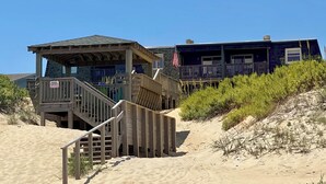 A beachfront property with a wooden staircase leading to a covered area, a flag, and a building with a balcony. - JL #R-10 | The Regency Condominium | Oceanfront | Community Pool (Kill Devil Hills)