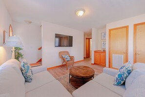 A living room with a sofa set, a wooden coffee table, a wicker chair, a TV mounted on the wall, and a door with a vent. - JL #309 | Sound Garden - Soundside - Sound Views - Private Heated Pool (Kitty Hawk)
