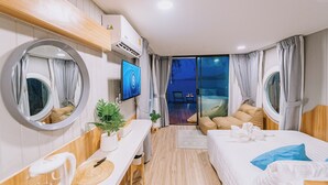 A modern hotel room with a wooden floor, a round mirror, a TV, a bed with pillows, a small table with a plant, and a view of the outdoors. - Sea Sand See Sky Beach Front Resort (Thep Krasattri)