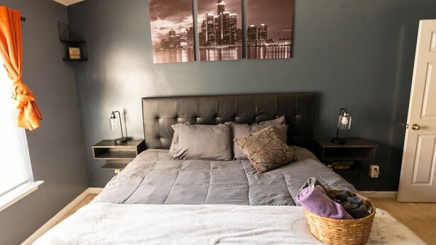 A bedroom with a large bed, a cityscape wall art, a nightstand, and a window with curtains.