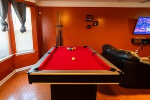 Une table de billard dans une pièce aux murs orange, avec un canapé en cuir et un téléviseur fixé au mur.