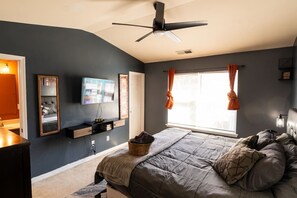 Une chambre à coucher comprenant un lit, un ventilateur de plafond, une télévision, une fenêtre avec des rideaux et une étagère fixée au mur.