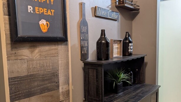 A wall with a framed sign saying 'EMPTY R. REPEAT' and a wine cellar sign. There are two bottles on a shelf, a potted plant, and a framed picture. - Downtown Atlanta 3BR Condo • Near Stadium & Arena (Atlanta)