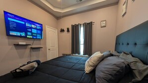 A bedroom with a bed, a television mounted on the wall, a window with curtains, and a wall-mounted shelf. - Cozy 4BR with Porch, King Bed Near Mercedez Benz & Downtown, Pet Friendly (Atlanta)