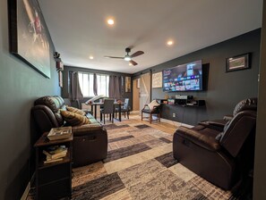 A living room with a sofa, a coffee table, a TV, a ceiling fan, and a dining area with a table and chairs. - Lux 2BR Condo w/ King Beds DTWN (Atlanta)
