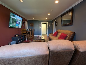 Ein Wohnzimmer mit einer Couch, einem Hocker, einem Fernseher und einem Essbereich mit Stühlen und einem Tisch.