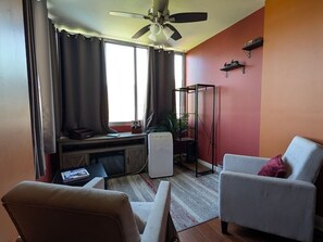 Ein Wohnzimmer mit Deckenventilator, einer Couch, einem Sessel, einem kleinen Tisch, einem Fernseher und einem Fenster mit Vorhängen.