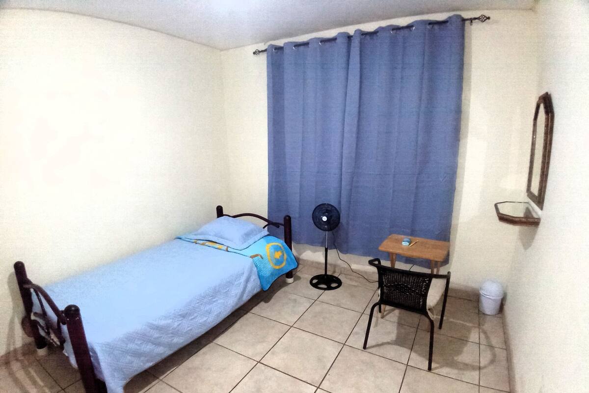 A small room with a bed, a desk, a chair, a fan, a mirror, and a curtain.