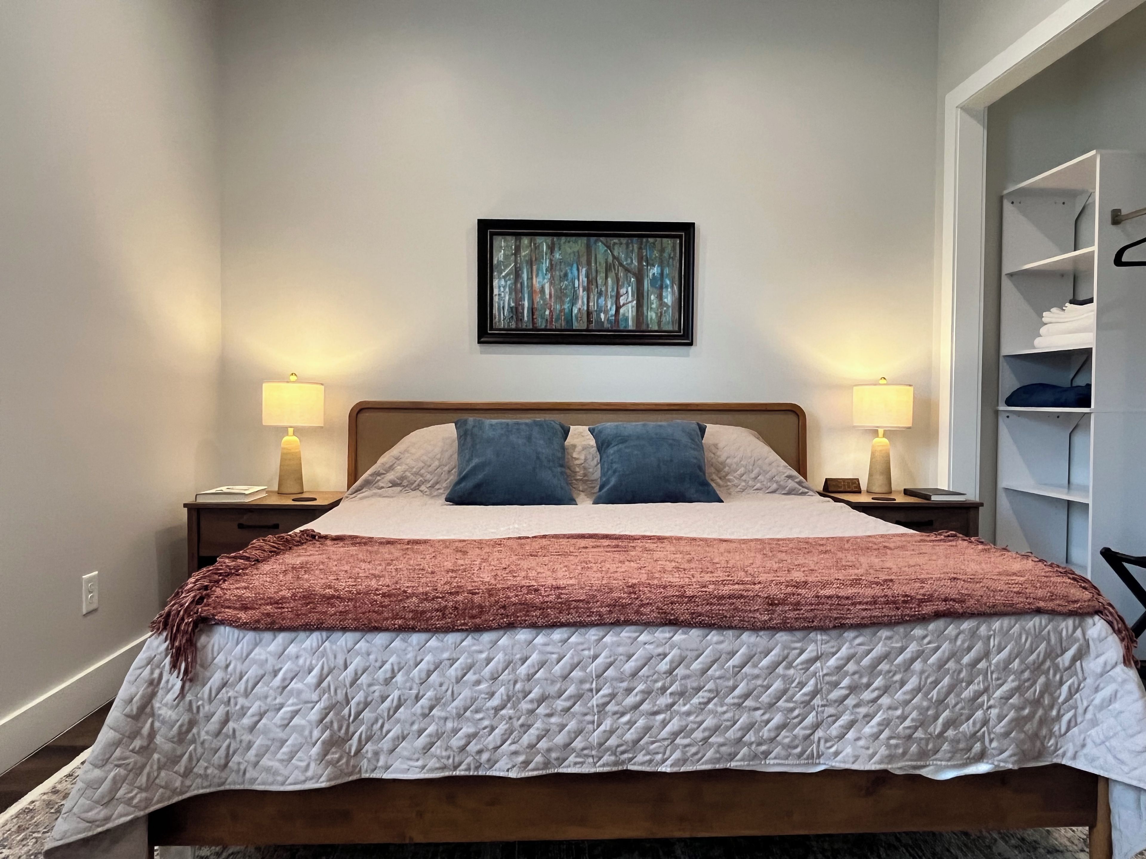 A neatly made bed with a quilted cover, two bedside lamps, a framed artwork above the bed, and a closet with shelves to the right.
