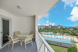 Ein Balkon mit Tisch und Stühlen, der auf einen Poolbereich mit Palmen und einem Gebäude im Hintergrund blickt.