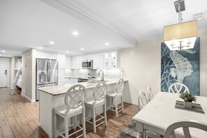 Eine moderne Küche mit weißen Schränken, einem Edelstahlkühlschrank, einer Mikrowelle und einem Spülbecken. Weiße Stühle stehen um einen Tisch mit einer Zierpflanze.