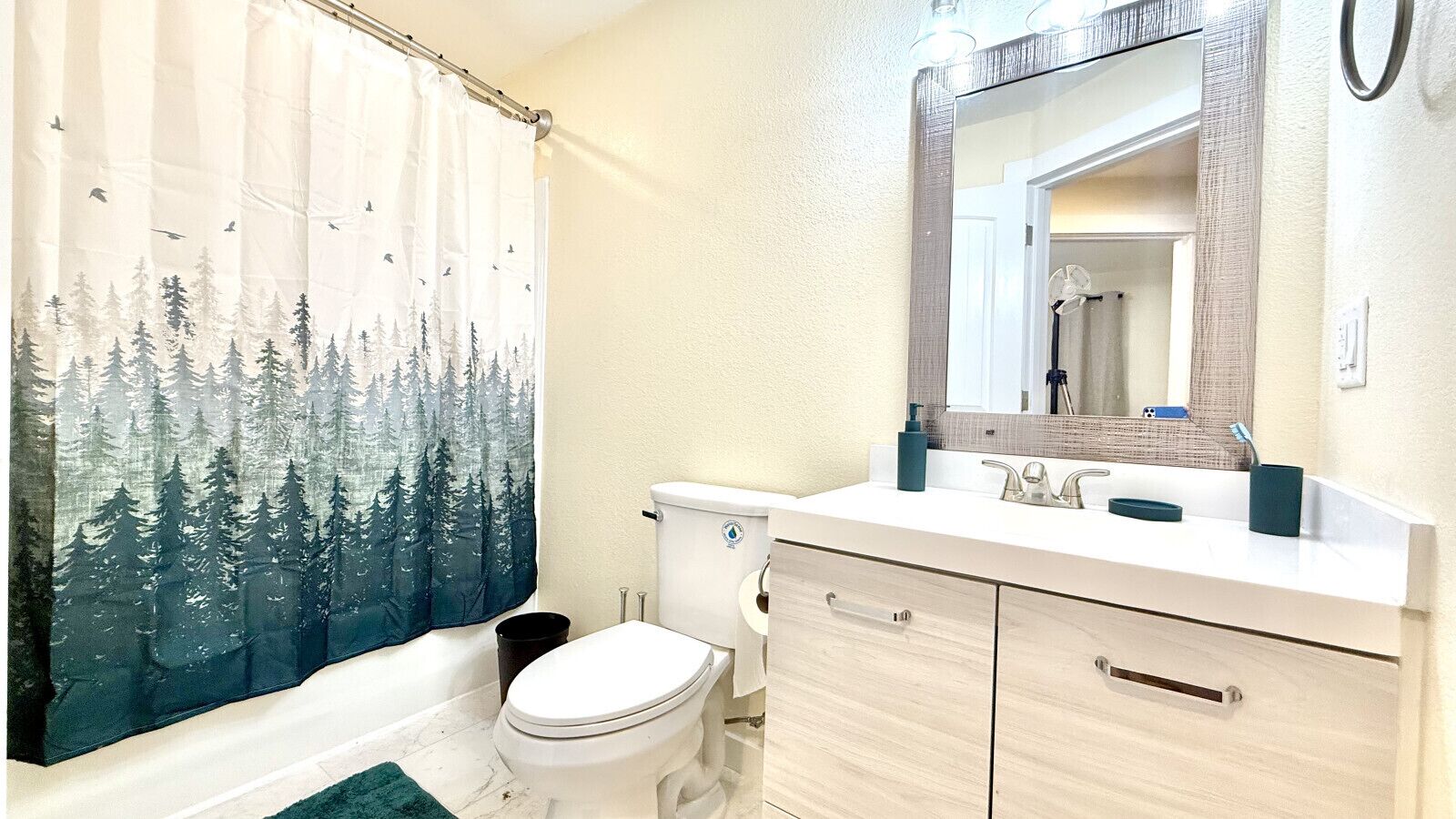 Une salle de bain avec un rideau de douche à motifs de forêt, des toilettes, un lavabo avec un miroir et une poubelle.