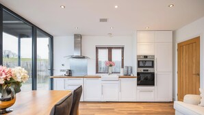 A modern kitchen with white cabinetry, a wooden dining table, and a large window with blinds. - Beautiful Eco Barn - sleeping 10 (Staveley)