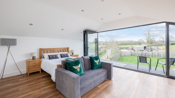 A bedroom with a bed, a grey sofa, a wooden nightstand, a floor lamp, and a large sliding glass door leading to an outdoor patio. - Beautiful Eco Barn - sleeping 10 (Staveley)