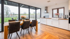 A modern kitchen with large windows, a dining area with a wooden table and chairs, and a view of the outdoors. - Beautiful Eco Barn - sleeping 10 (Staveley)