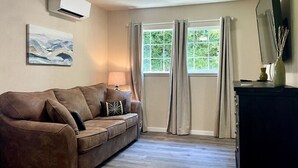 Uma sala de estar com um sofá marrom, uma luminária, um quadro, uma janela com cortinas e uma TV de tela plana em um suporte.