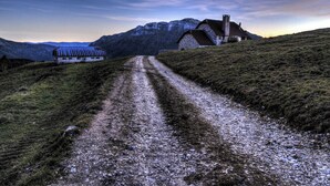 Un chemin de campagne menant à une maison aux murs en pierre et dotée d’une cheminée, entourée de montagnes.