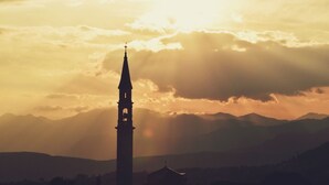 Le clocher d’une église se détache sur le ciel au coucher du soleil, avec des montagnes en arrière-plan.