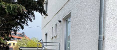 Un edificio residencial con una escalera de metal que conduce a una puerta, un sendero pavimentado y un árbol al fondo.