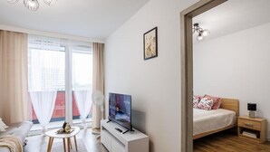 A modern hotel room with a flat-screen TV, a bed with a wooden headboard, a bedside table with a lamp, and a large window with sheer curtains. - Malila Apartments Łaszkiewicza 8 (Kraków)